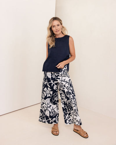 Woman wearing a navy sleeveless top and white floral pants against a white background