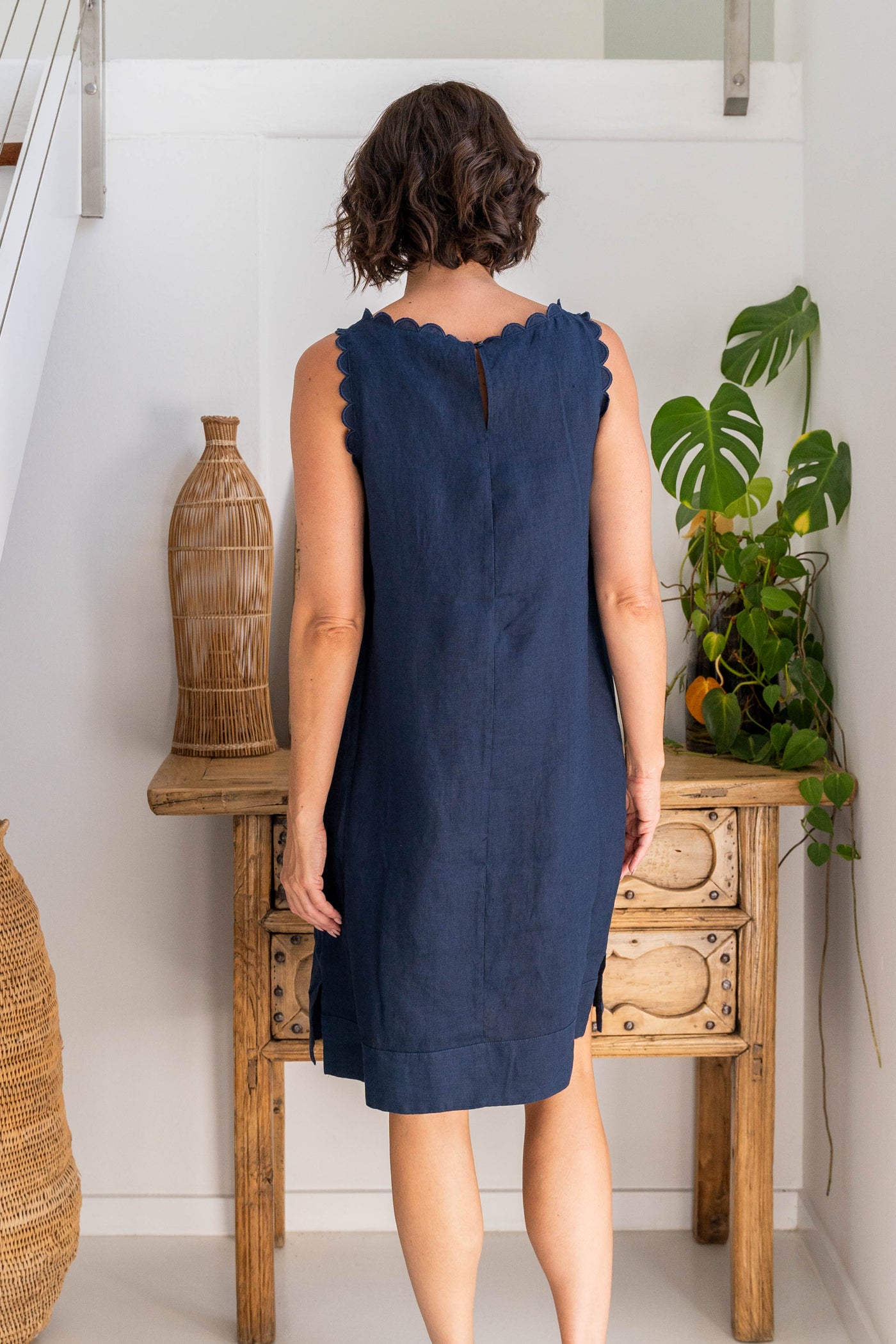 Woman wearing a blue dress standing in a room with wooden furniture and plants.
