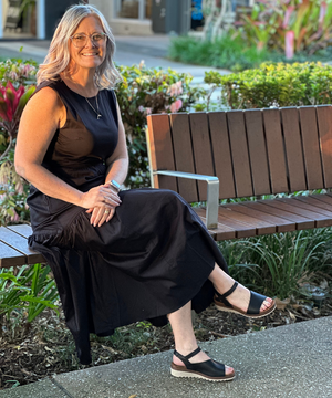 Woman in a black dress sitting on a wooden bench outdoors with plants and flowers in the background.